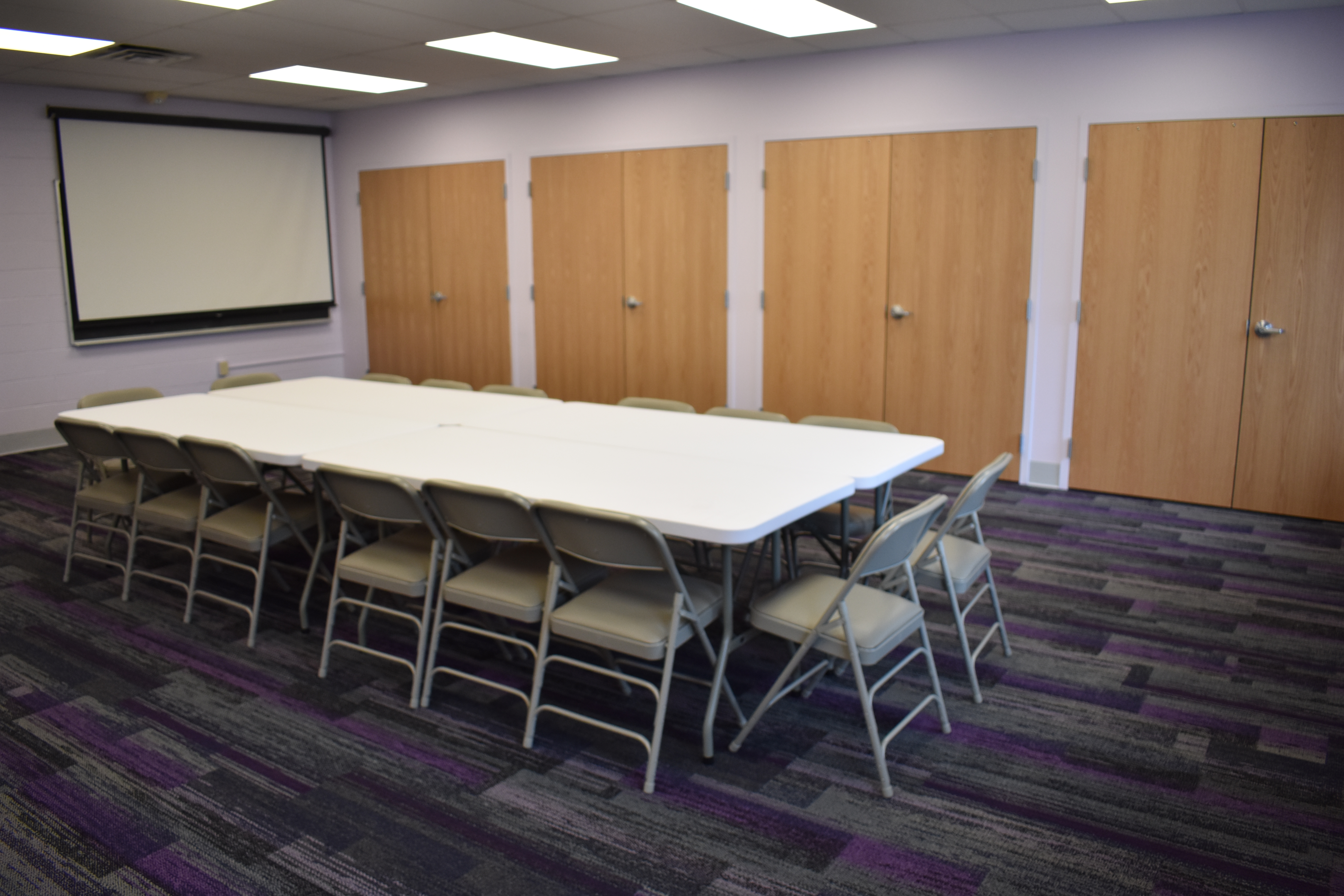 Photo of Community Room with tables and chairs, closet doors, and projector screen.