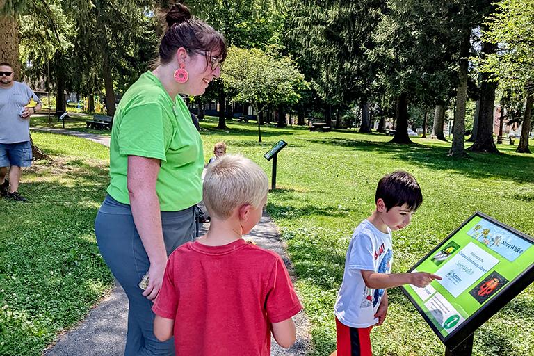 Two young boys stand beside library staff member as they read a StoryWalk page in the park.