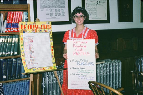 Smiling lady librarian holding Summer Reading Club poster